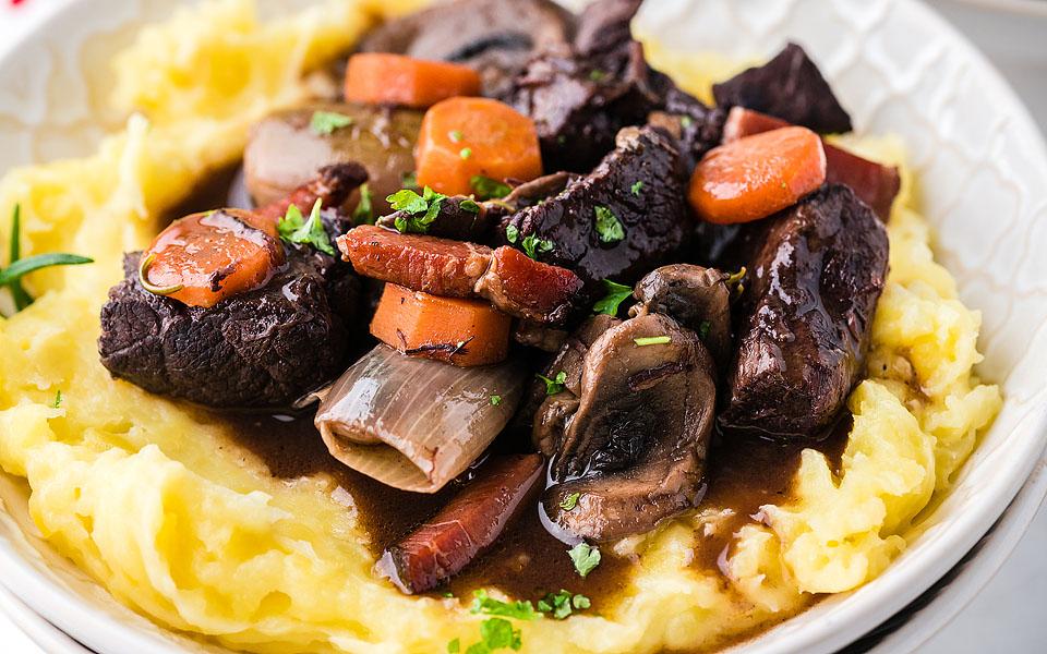 A plate of mashed potatoes topped with beef stew, mushrooms, carrots, onions, and garnished with chopped herbs.