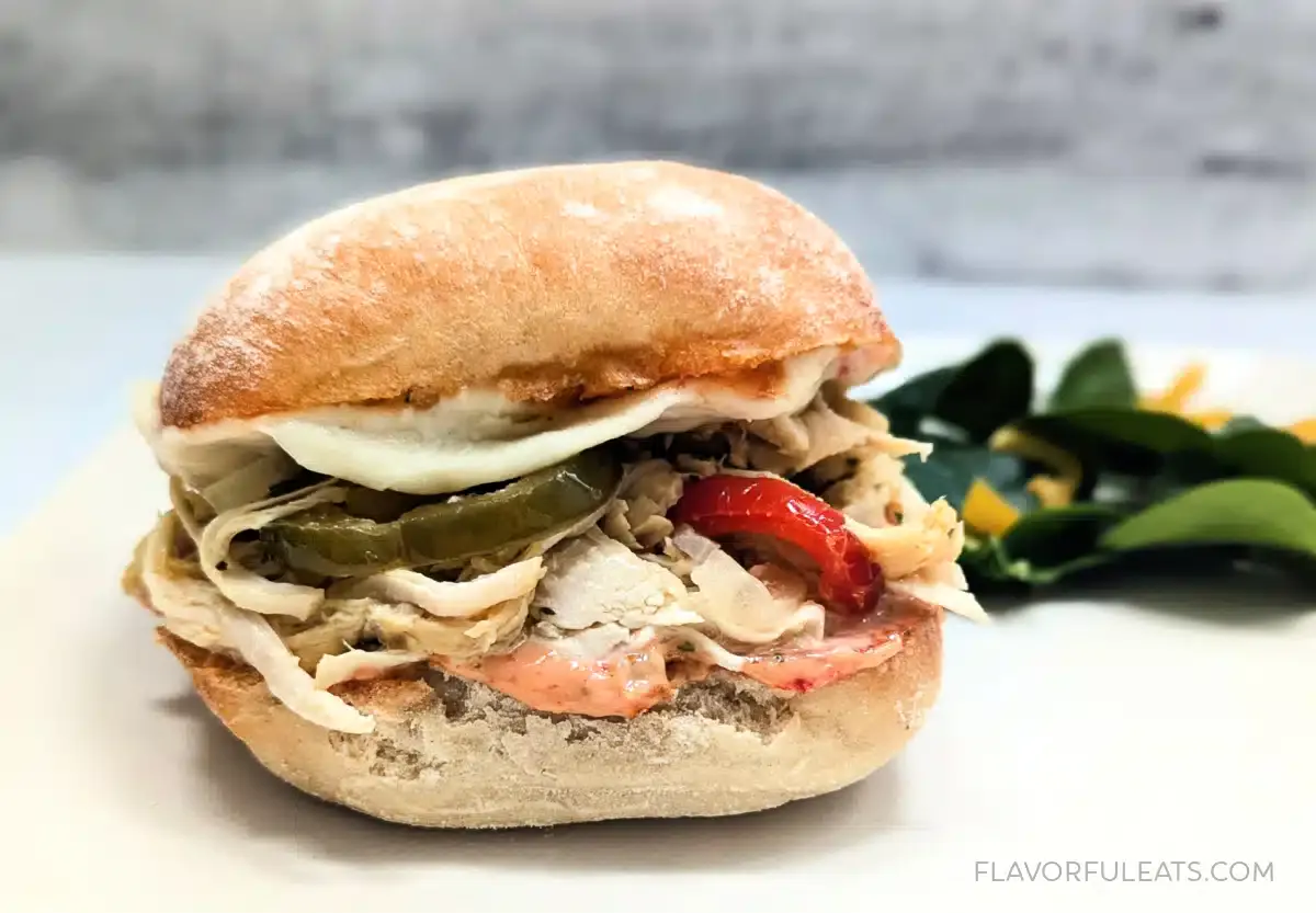 A shredded chicken sandwich with sliced pickles, red peppers, and a white bun, served on a white plate with leafy greens and orange slices in the background.