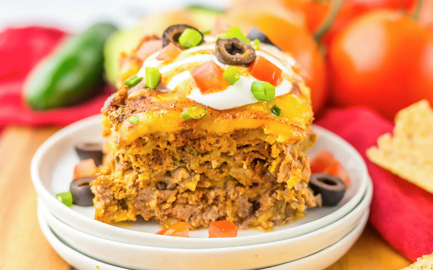 A slice of layered taco casserole topped with sour cream, chopped tomatoes, green onions, and sliced black olives, served on a white plate with tomatoes and chips in the background.
