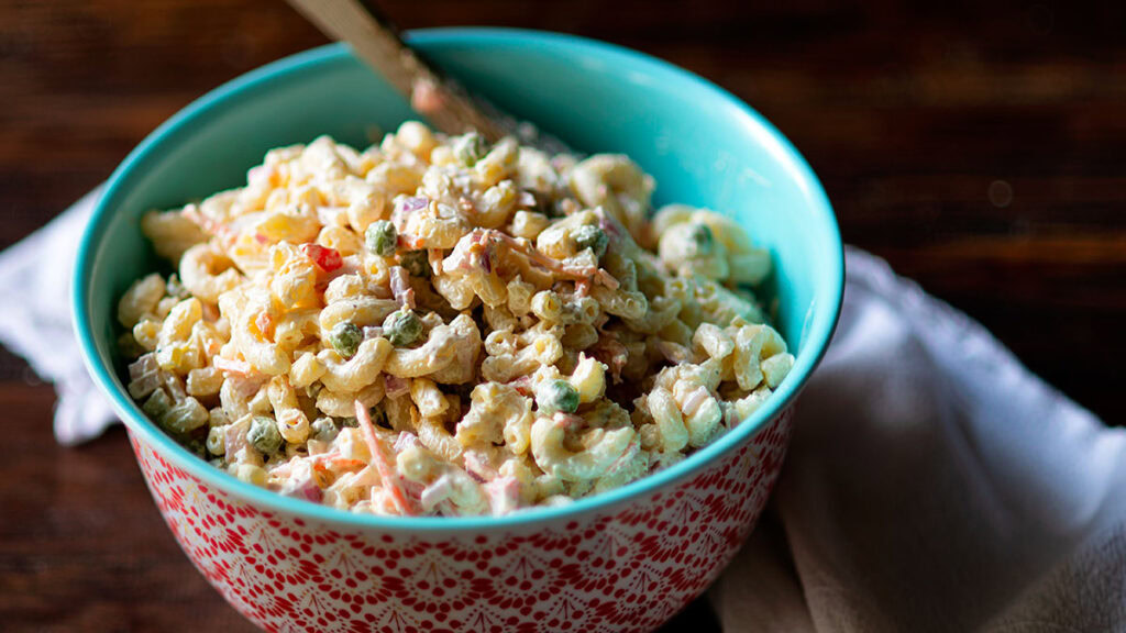A bowl of macaroni salad with vegetables and dressing, placed on a white cloth with a spoon inside the bowl.
