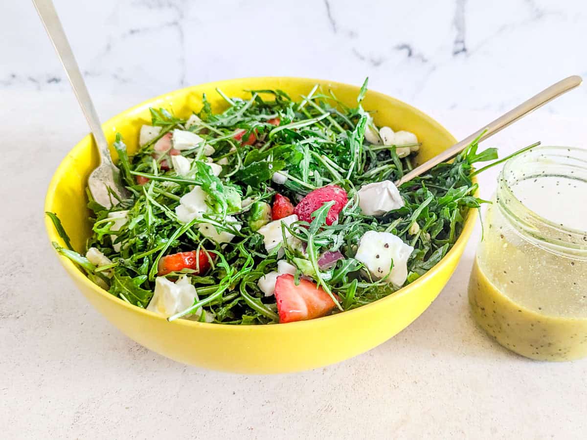 A yellow bowl filled with arugula salad, strawberries, red onion, and mozzarella, with two spoons and a jar of salad dressing beside it.