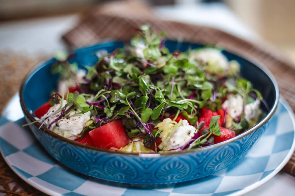 A blue bowl filled with a salad featuring mixed greens, watermelon cubes, and dollops of cheese on a patterned plate.