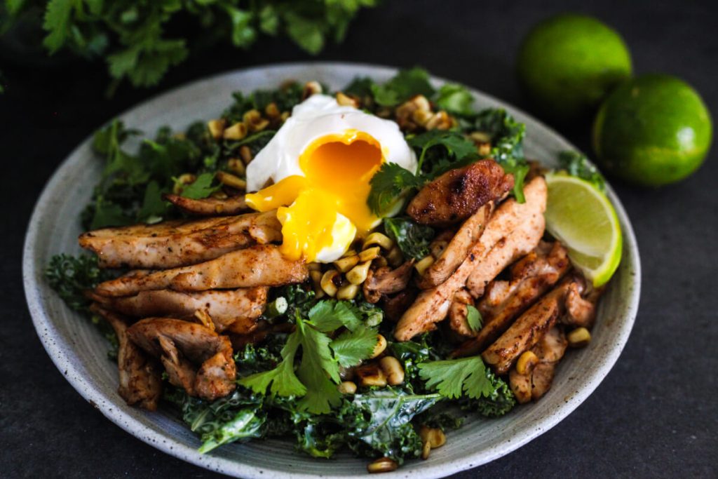 A plate of grilled chicken strips on a bed of leafy greens, topped with a poached egg and garnished with cilantro, seeds, and a lime wedge. Two limes are in the background.