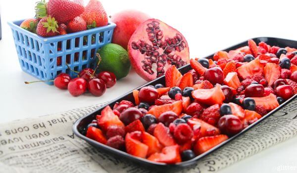 A tray filled with assorted fresh berries, surrounded by a basket of strawberries, a halved pomegranate, a lime, and cherries on a newspaper-covered surface.