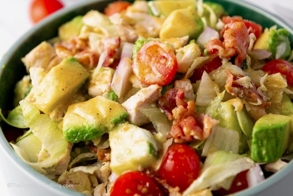 A close-up of a salad with avocado, cherry tomatoes, lettuce, bacon, and red onion in a green bowl.