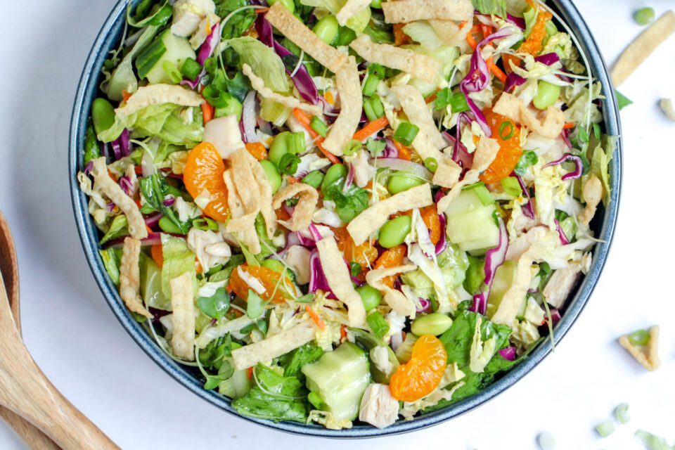 A bowl of salad with lettuce, shredded cabbage, cucumber, mandarin orange slices, edamame, and crispy wonton strips.