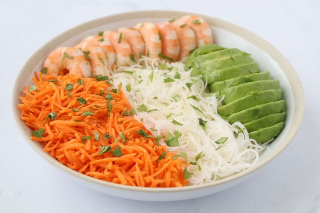 A bowl with sliced shrimp, avocado, shredded carrots, and rice noodles, garnished with chopped herbs on top.
