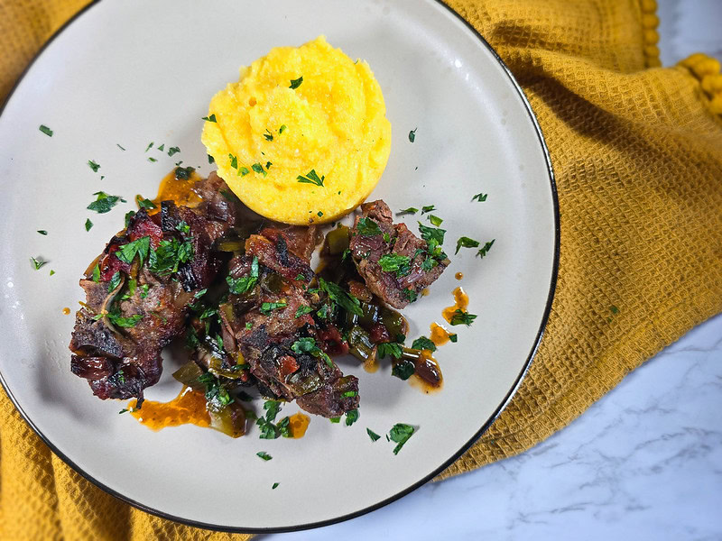 A plate with three pieces of herb-garnished braised meat and a round serving of polenta, set on a yellow textured cloth.