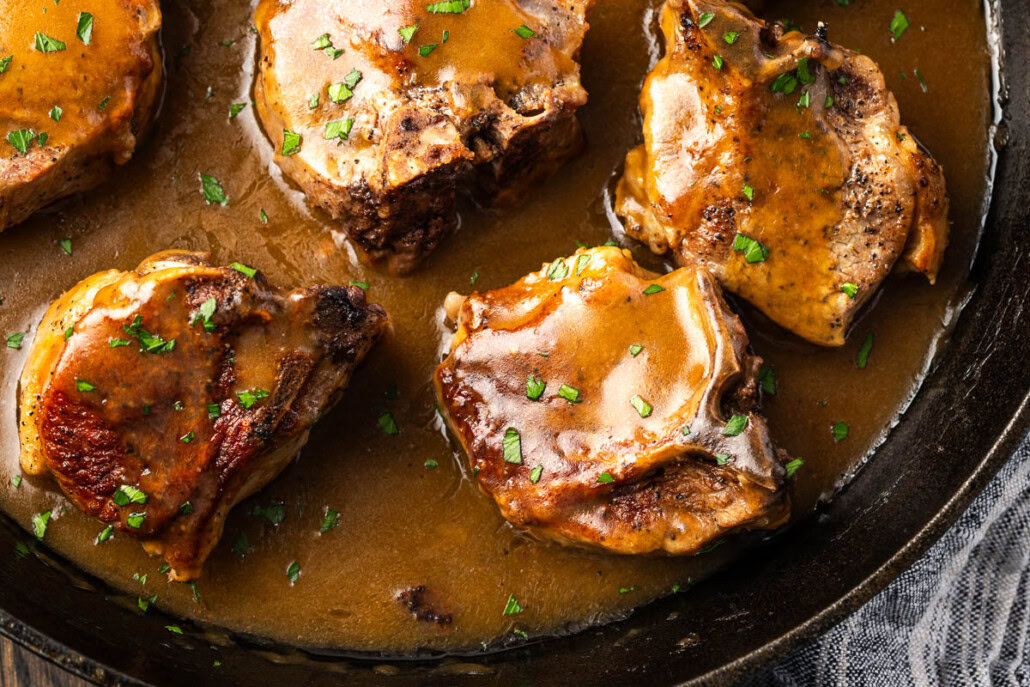 Four cooked pork chops covered in brown gravy, garnished with chopped herbs, served in a black skillet.