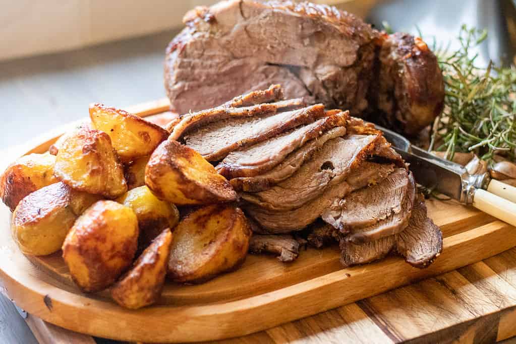 Sliced roast lamb and golden roasted potatoes arranged on a wooden cutting board with a knife and sprigs of rosemary.