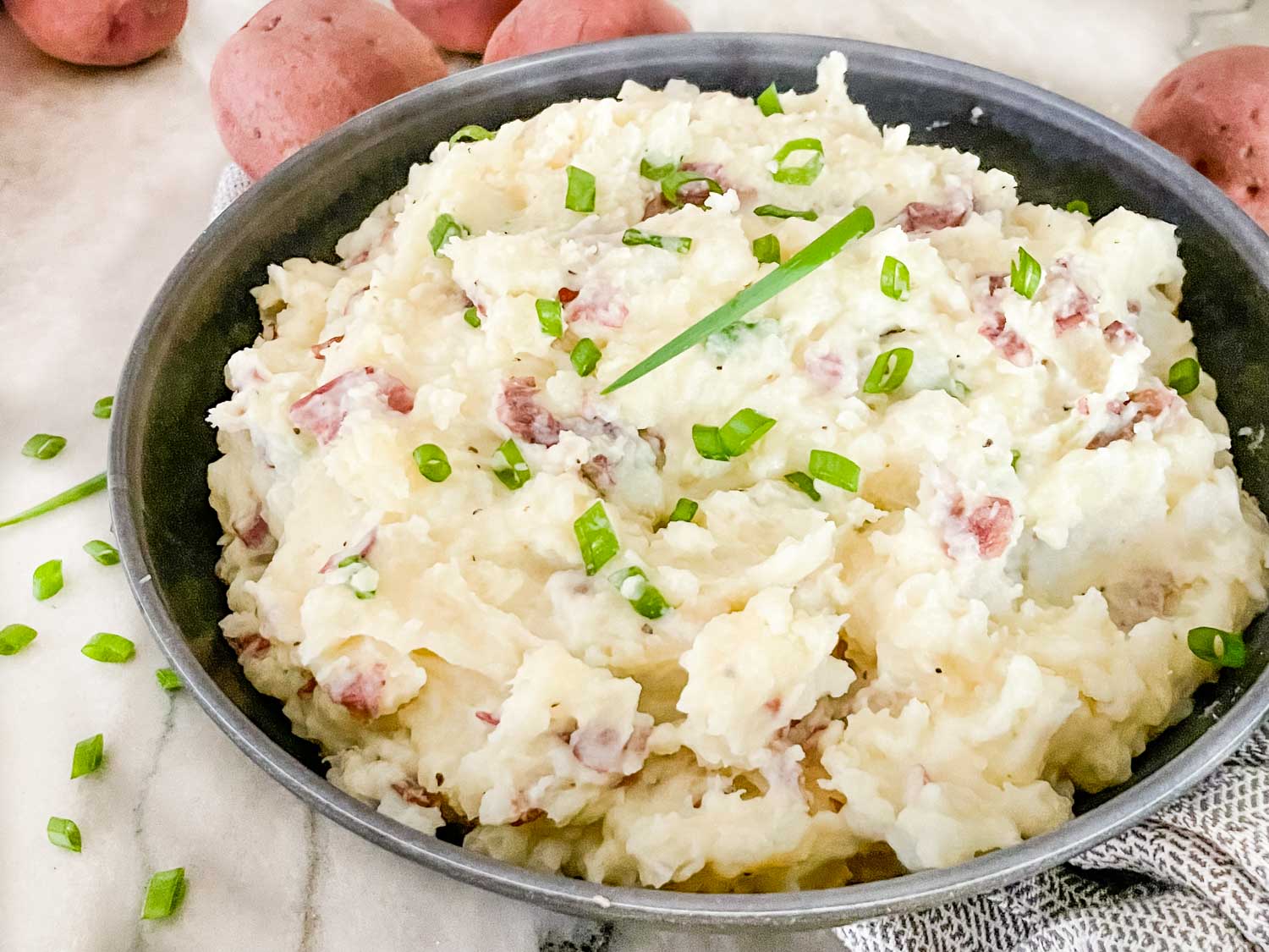 A bowl of mashed potatoes with red potato skin pieces, garnished with chopped green onions, sits on a marble surface with whole red potatoes in the background.