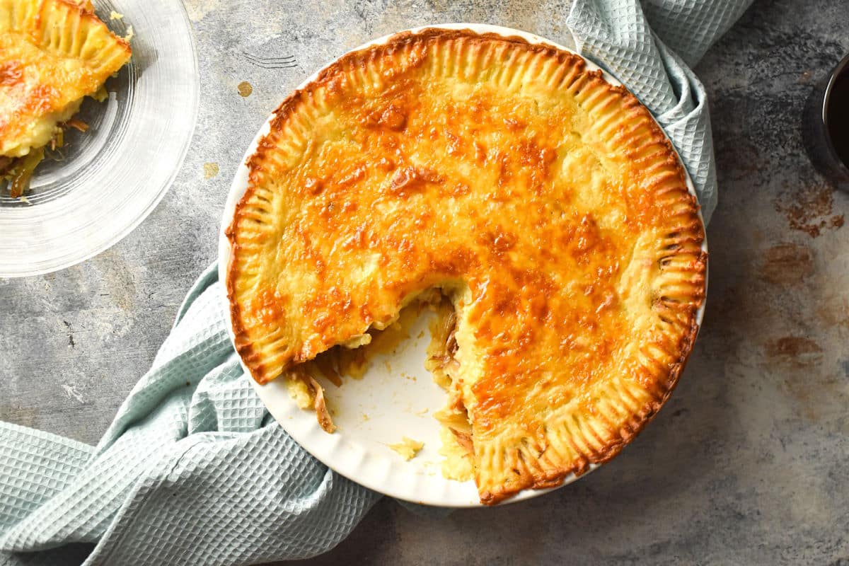 A golden-brown pie with a slice missing sits in a white dish on a gray surface, next to a blue textured cloth.