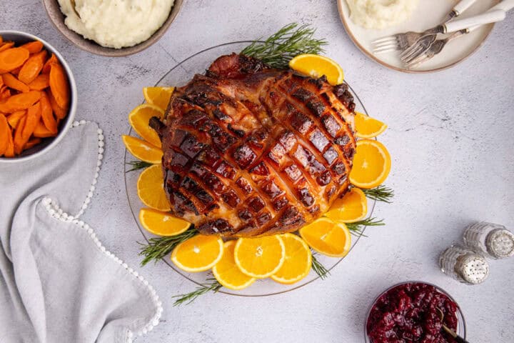 A glazed ham garnished with orange slices and rosemary sits on a plate, surrounded by bowls of carrots, mashed potatoes, and cranberry sauce on a light table.