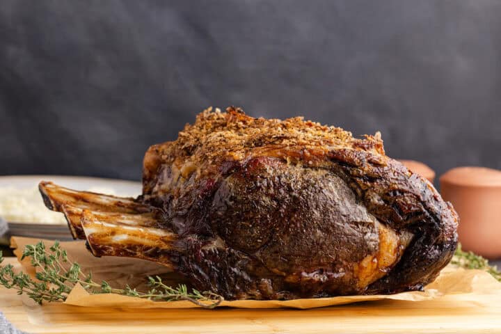 A cooked bone-in prime rib roast with a seasoned crust rests on a wooden board, garnished with sprigs of fresh thyme.
