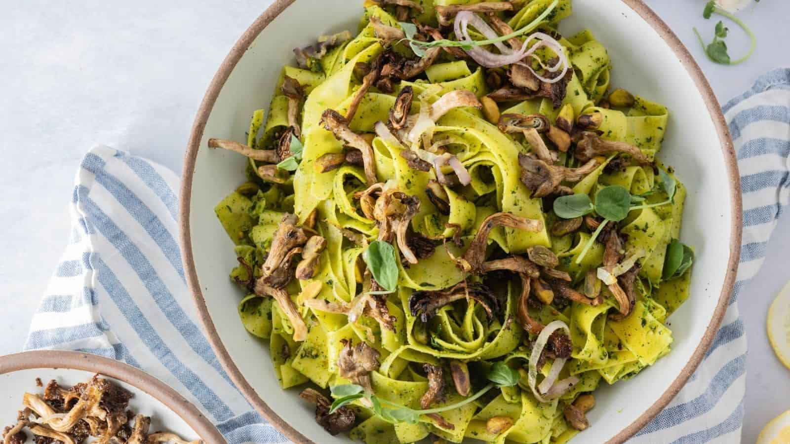 A bowl of wide green pasta ribbons mixed with sautรฉed mushrooms, fresh herbs, and thinly sliced onions sits on a striped cloth.