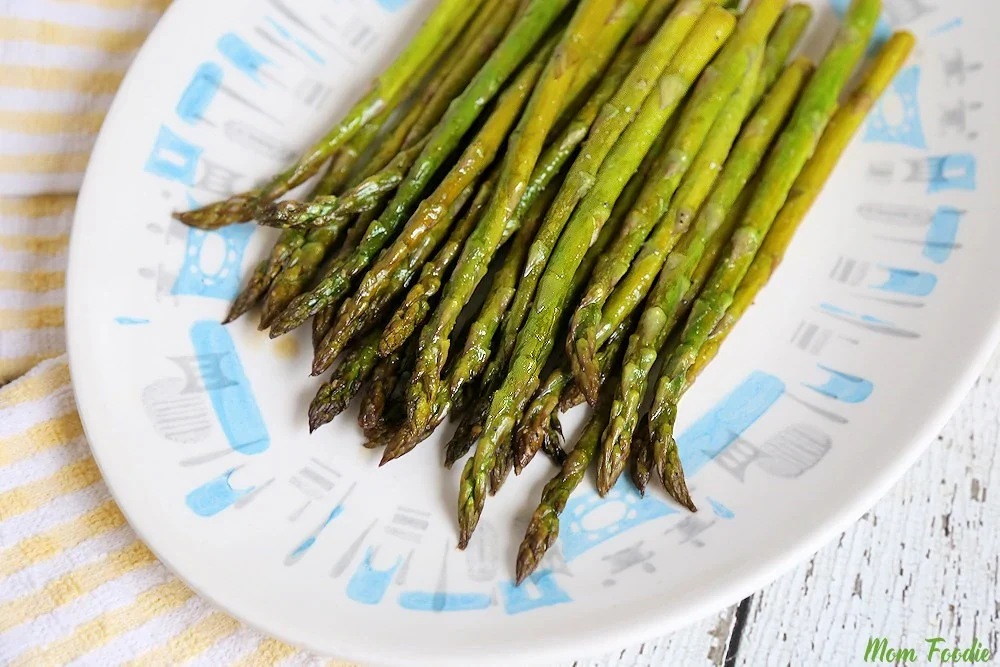 A plate of cooked asparagus spears arranged in a row on a white plate with a blue pattern, placed on a striped cloth next to a white wooden surface.