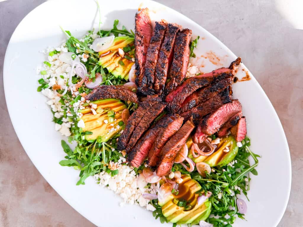 Sliced grilled steak served on a bed of arugula with avocado, feta cheese, shallots, and nuts on a white oval plate.