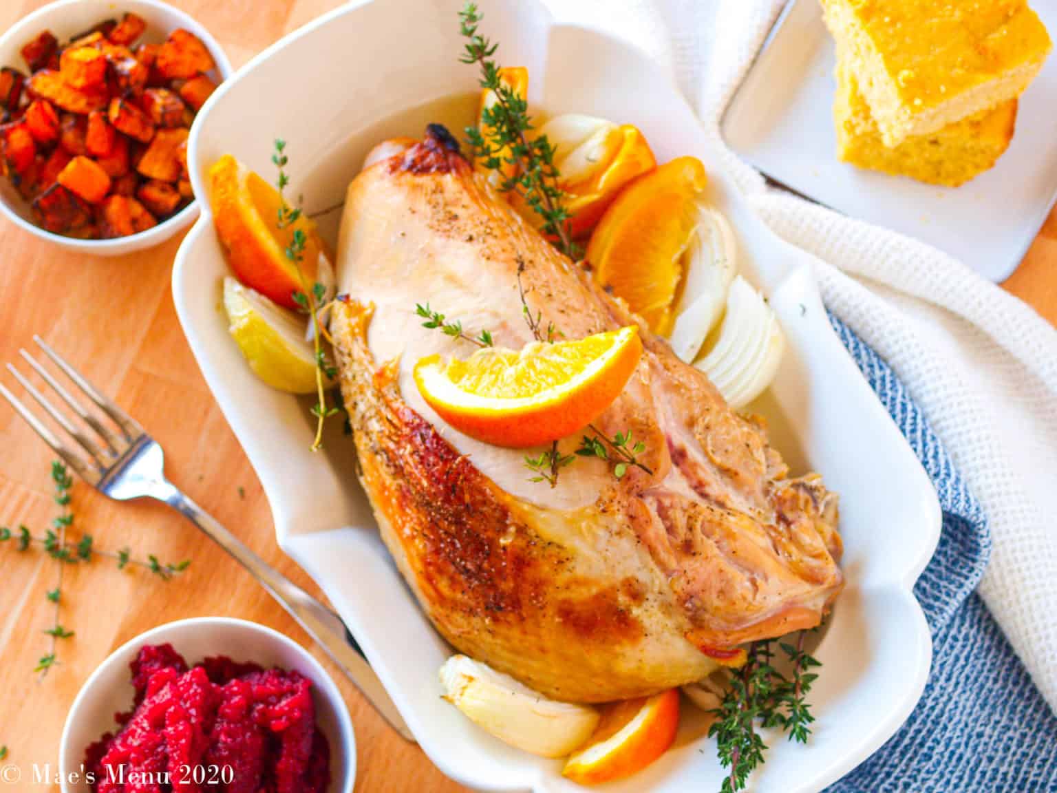 A roasted turkey breast garnished with orange slices and herbs is served in a white dish, with bowls of cranberry sauce, roasted vegetables, and cornbread on the side.
