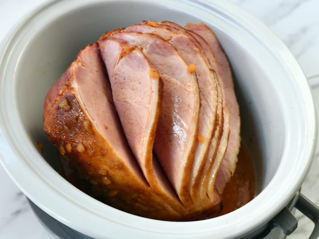 A sliced ham sits in a white slow cooker with some juices at the bottom, ready for cooking.