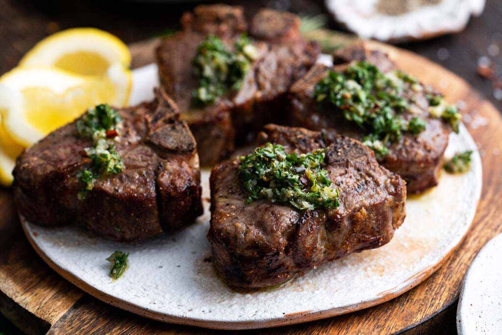 Four grilled lamb chops topped with green herb sauce are served on a white plate, with lemon wedges on the side.