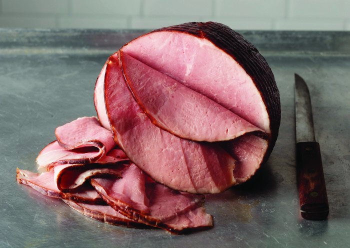 A large piece of sliced ham sits on a metal surface next to a carving knife, with several thin slices arranged in front.