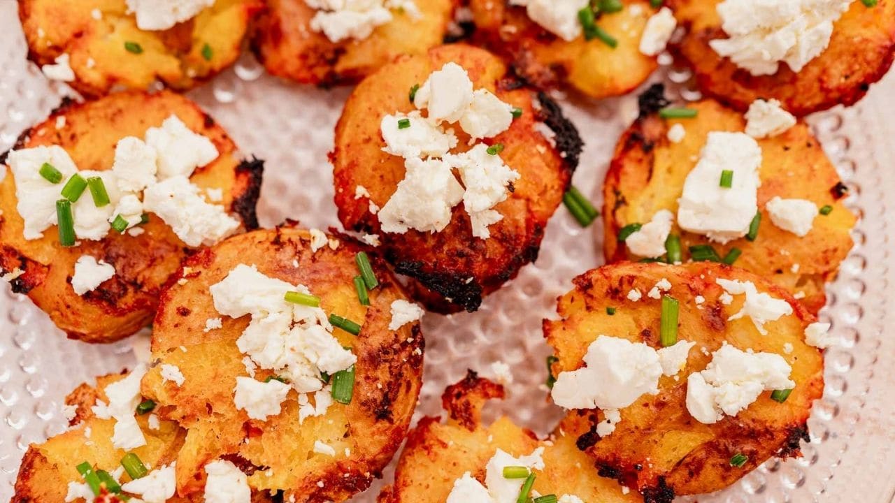 Close-up of smashed roasted potatoes topped with crumbled white cheese and chopped chives on a textured glass plate.