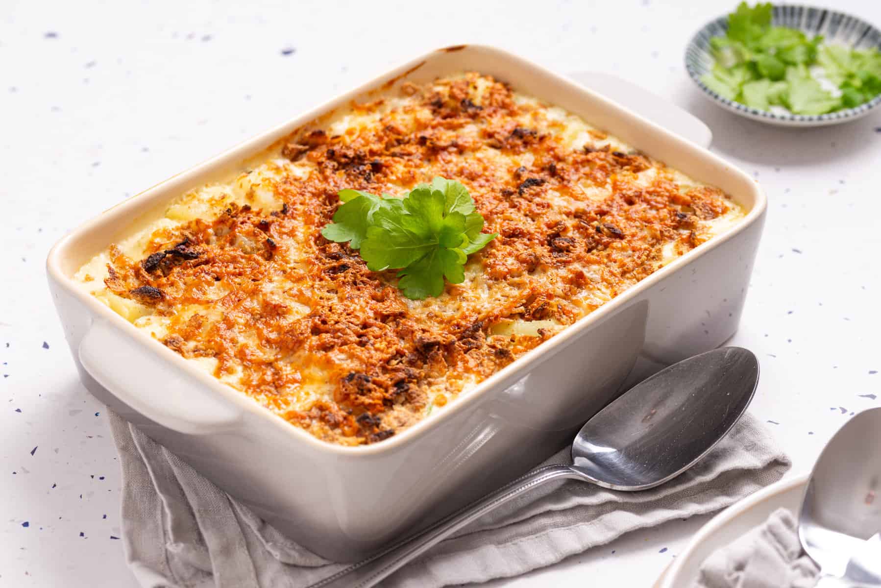 A baked casserole topped with melted cheese and garnished with fresh parsley, placed in a white rectangular dish beside a spoon and fork.