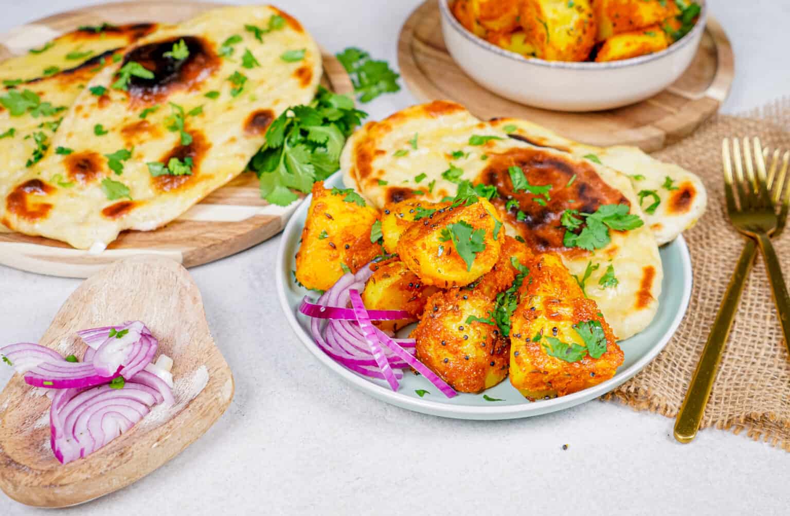 A plate with seasoned paneer tikka garnished with cilantro, sliced red onions, and naan bread, with more naan and paneer tikka in the background.