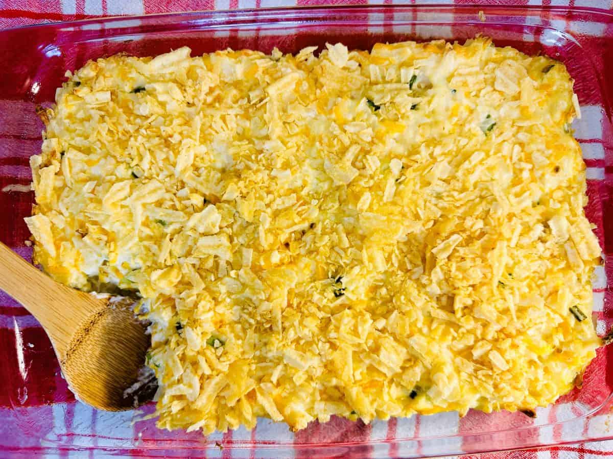 A glass baking dish of casserole with a golden, crumbly topping. A wooden spoon rests in a corner where a portion has been served.