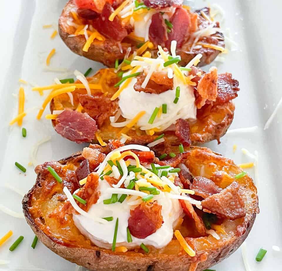 Three loaded potato skins topped with sour cream, shredded cheese, bacon pieces, and chopped chives on a white plate.