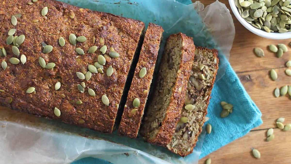 A loaf of seed-topped bread, partially sliced, sits on parchment and a blue cloth next to a bowl of pumpkin seeds.
