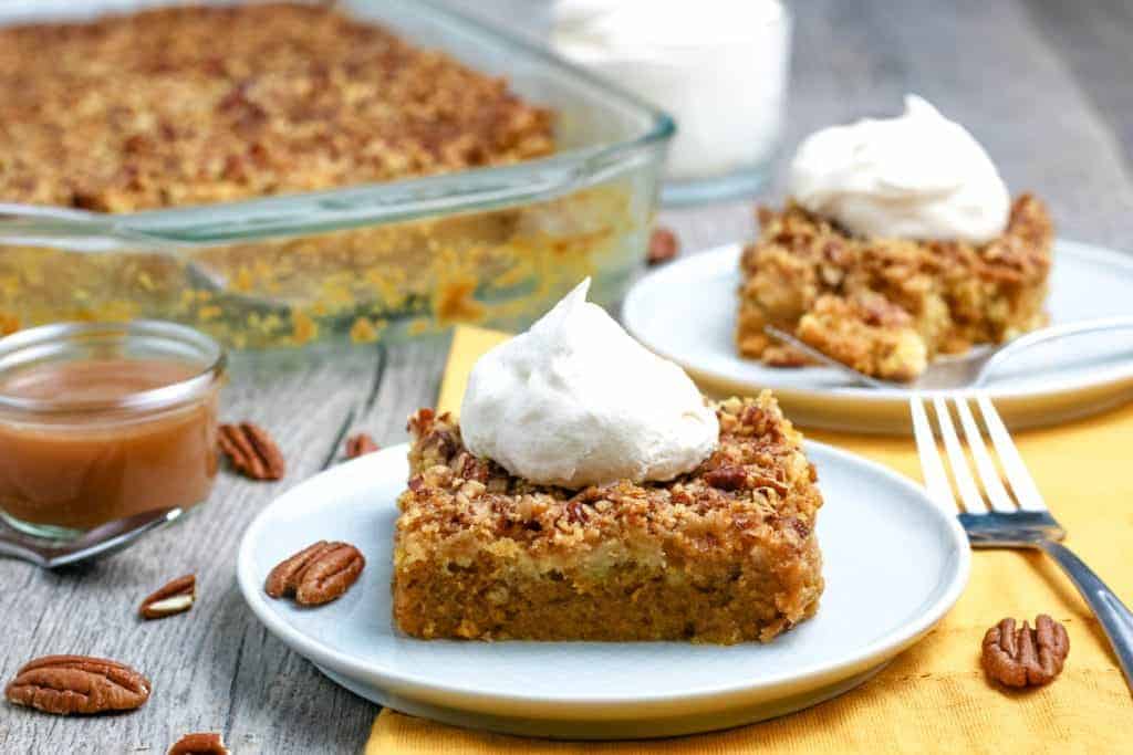 A slice of pumpkin dessert topped with whipped cream on a plate, with a baking dish of the same dessert, caramel sauce, and pecans in the background.