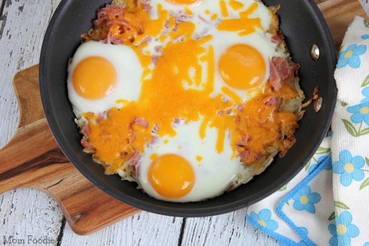 A skillet with four sunny-side up eggs, melted cheddar cheese, and shredded ham on a wooden cutting board next to a floral-patterned napkin.