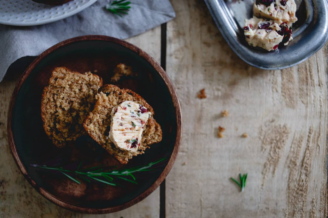 A wooden bowl with two slices of bread, one topped with herb and cranberry butter. A metal dish with more butter is visible in the corner, with rosemary sprigs on the table.