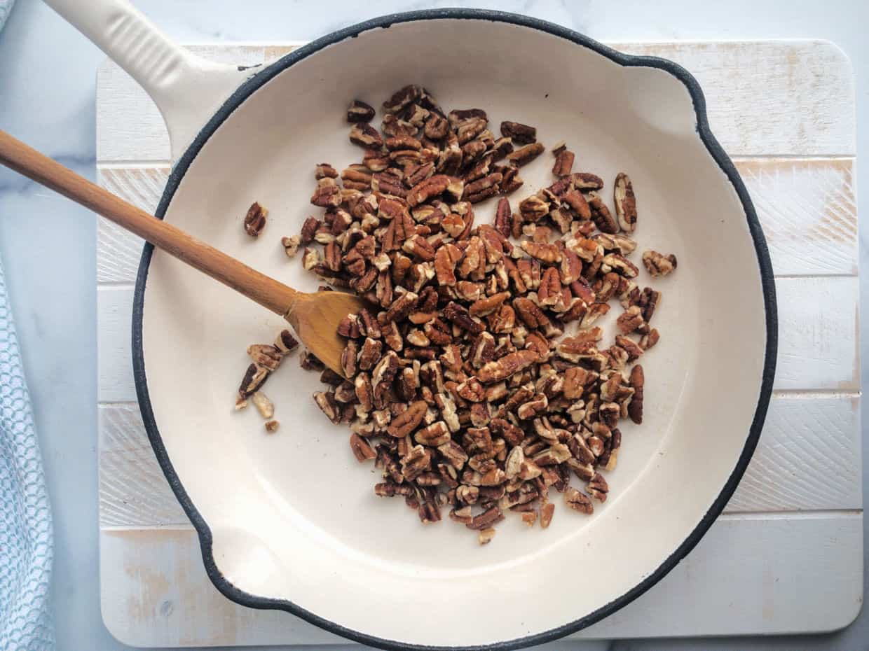 Chopped pecans being stirred with a wooden spoon in a white skillet on a white wooden surface.