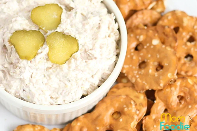A white bowl of creamy dip topped with three pickle slices is next to a pile of pretzel crisps.