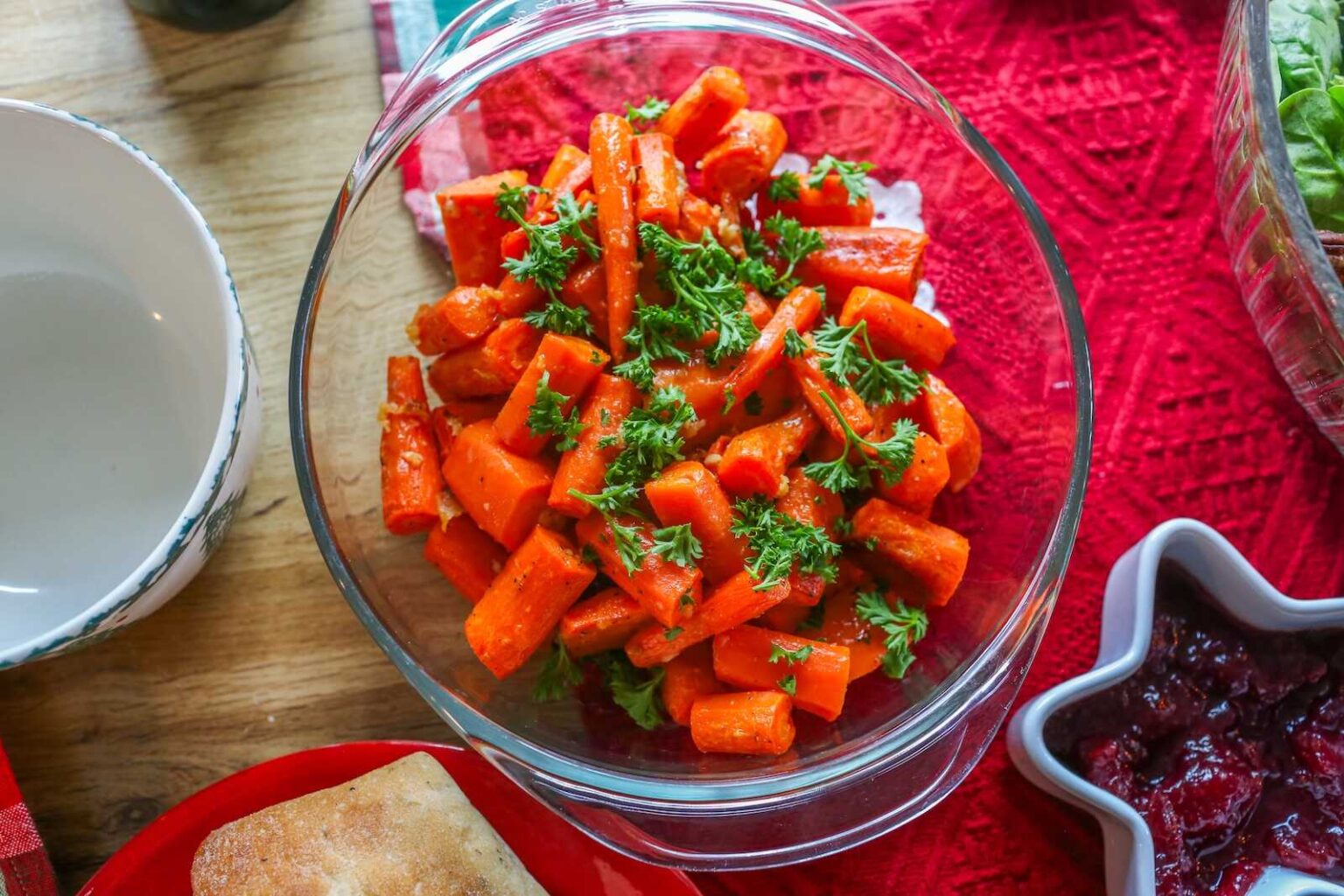 A glass bowl filled with cooked carrot sticks garnished with chopped parsley, placed on a red tablecloth next to other dishes.