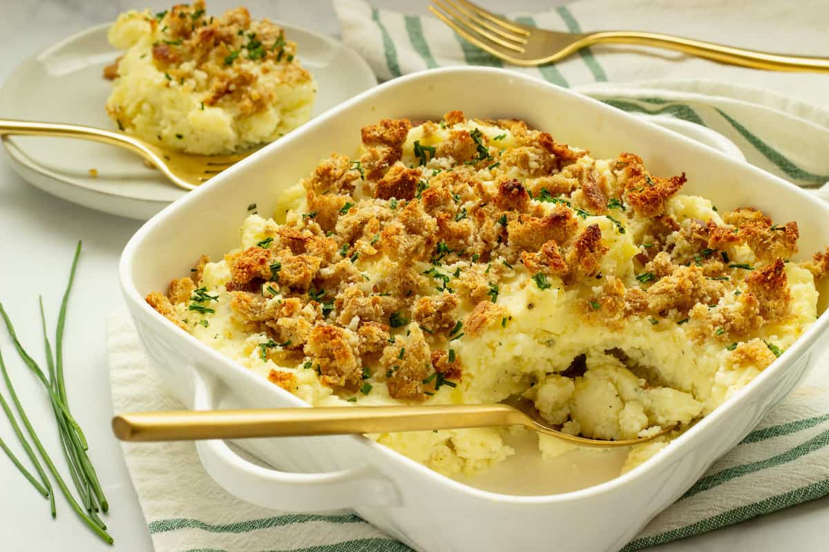 A white baking dish filled with baked mashed potatoes topped with golden breadcrumbs, with a serving spoon and a portion served on a plate in the background.