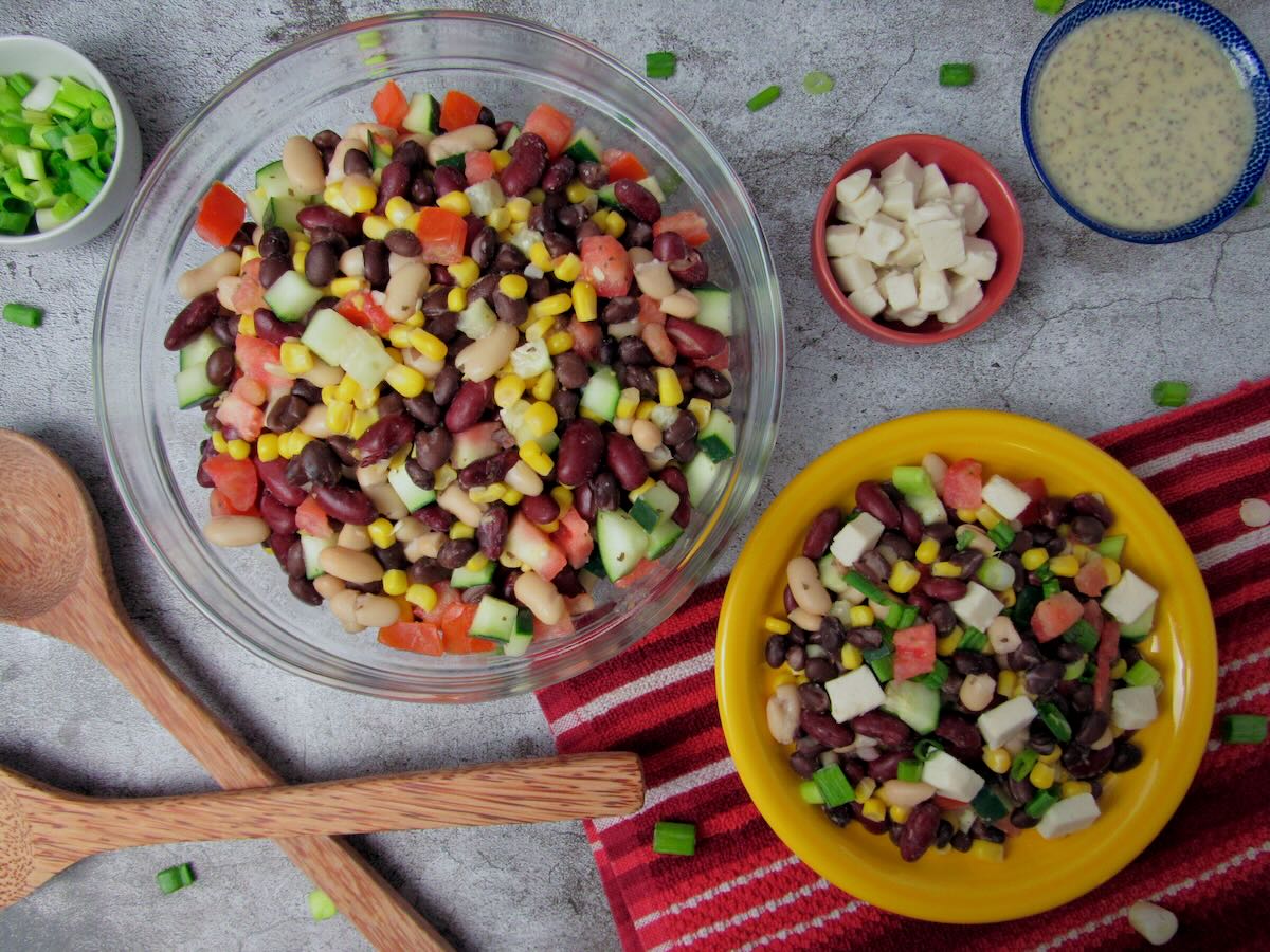 A bowl and a plate of mixed bean salad with corn, cucumber, and diced vegetables, surrounded by salad dressing, cheese cubes, green onions, and wooden utensils on a red cloth.