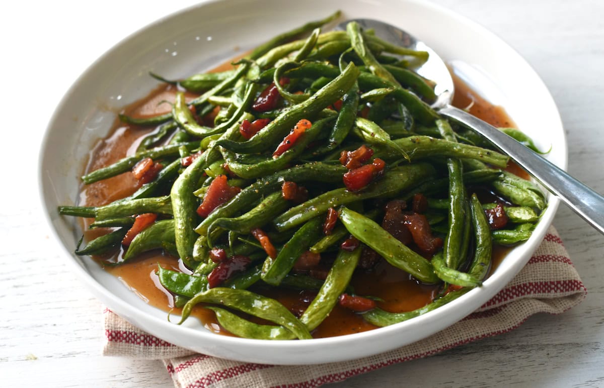 A white plate of cooked green beans mixed with pieces of bacon in a reddish-brown sauce, with a spoon on the side.