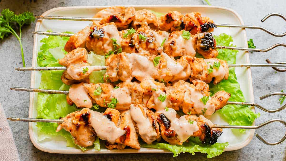 Four grilled chicken skewers with creamy sauce are arranged on a rectangular plate lined with lettuce leaves, garnished with chopped herbs.