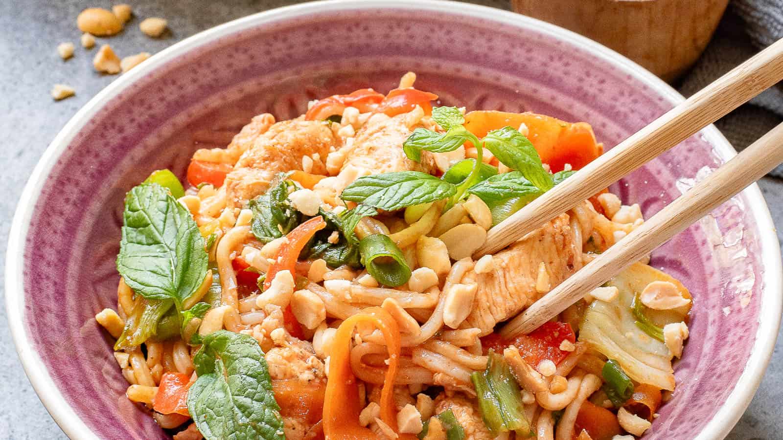 A bowl of noodle stir-fry with vegetables, chicken, chopped peanuts, and fresh mint leaves, served with chopsticks.