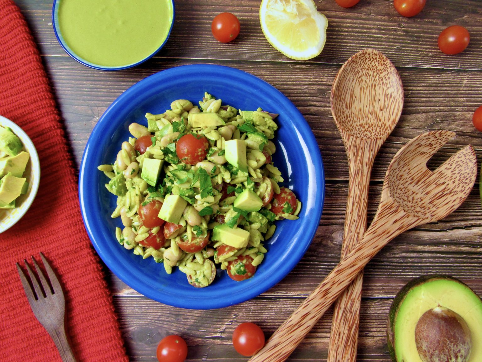 A blue plate with pasta salad containing avocado, cherry tomatoes, and greens sits on a wooden table with wooden utensils, a halved avocado, lemon, and a bowl of green dressing nearby.