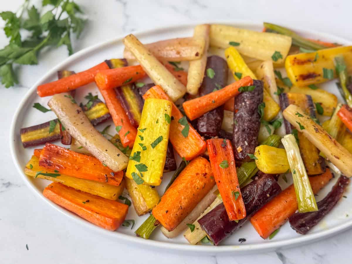 A white plate with roasted multicolored carrots and parsnips, garnished with chopped parsley, on a white surface.