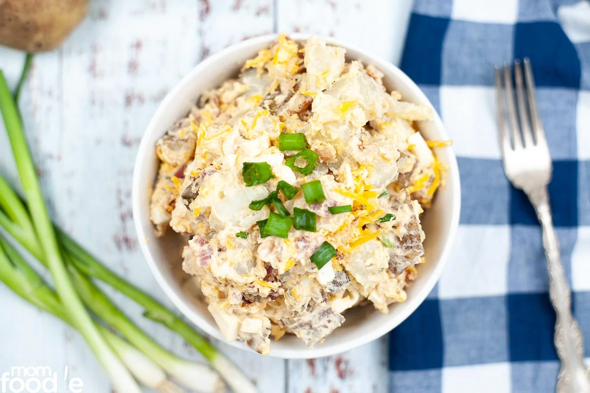 A bowl of potato salad topped with chopped green onions, placed on a light wooden surface next to a fork, green onions, and a blue plaid napkin.