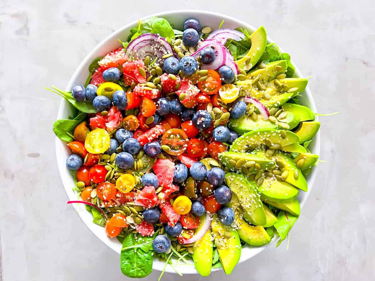 A bowl of salad with spinach, avocado slices, cherry tomatoes, blueberries, red onion, grapefruit, pumpkin seeds, and a sprinkle of sesame seeds.