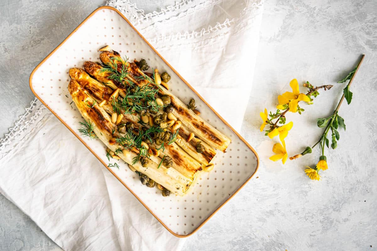 A rectangular plate with cooked white asparagus spears topped with herbs, capers, and pine nuts, placed on a white cloth next to yellow flowers.