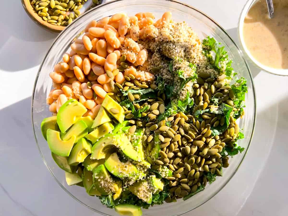 A glass bowl filled with chopped kale, avocado slices, white beans, pepitas, and a sprinkling of hemp seeds, with a bowl of dressing nearby.