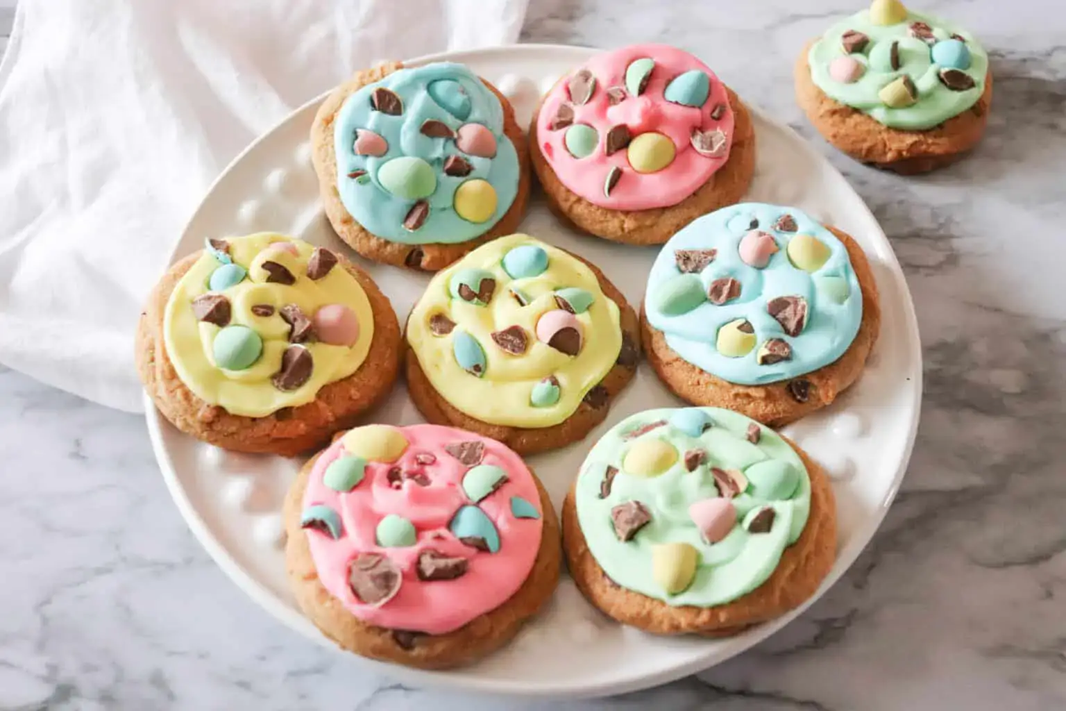 A white plate holds eight cookies topped with pastel-colored icing and decorated with chocolate pieces and candy-coated chocolates.