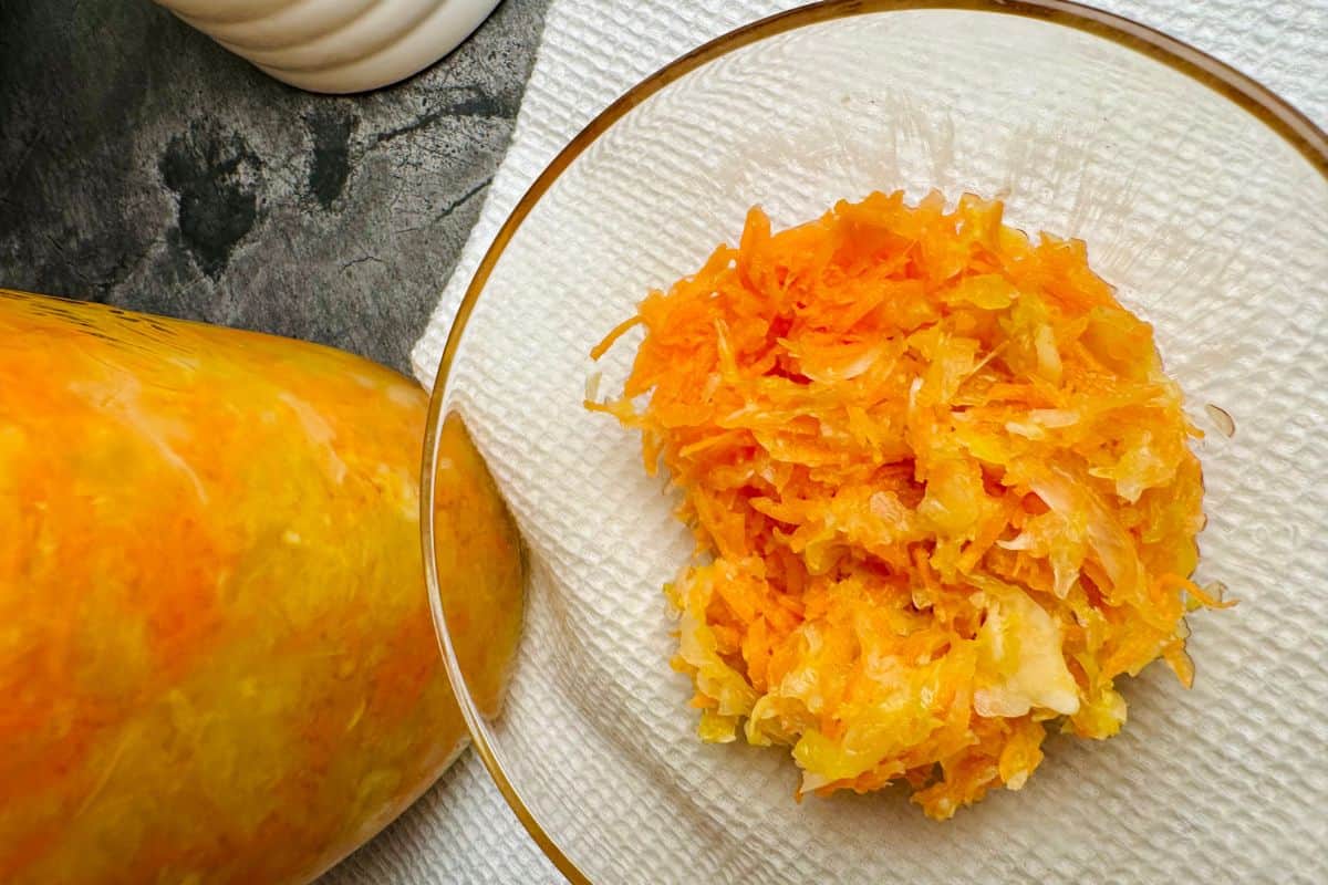 A bowl of shredded orange and white vegetables, likely fermented, sits on a paper towel next to a jar filled with a similar mixture.